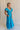 Full body side view of female model wearing the Gianna Ruffle Short Sleeve Tiered Maxi Dress in Blue which features Lightweight Fabric, Tiered Body, Midi Length, Pockets on each side, V-Neckline with a Tie Detail and Ruffle Short Sleeves. the dress is available in pink, aqua blue and white.