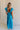 Full body back view of female model wearing the Gianna Ruffle Short Sleeve Tiered Maxi Dress in Blue which features Lightweight Fabric, Tiered Body, Midi Length, Pockets on each side, V-Neckline with a Tie Detail and Ruffle Short Sleeves. the dress is available in pink, aqua blue and white.