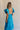 Back view of female model wearing the Gianna Ruffle Short Sleeve Tiered Maxi Dress in Blue which features Lightweight Fabric, Tiered Body, Midi Length, Pockets on each side, V-Neckline with a Tie Detail and Ruffle Short Sleeves. the dress is available in pink, aqua blue and white.