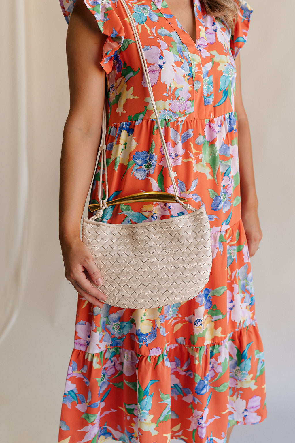 Model is wearing the Nadia Woven Purse on her shoulder. It features a cream woven body topped with a gold bar accent and cream straps.