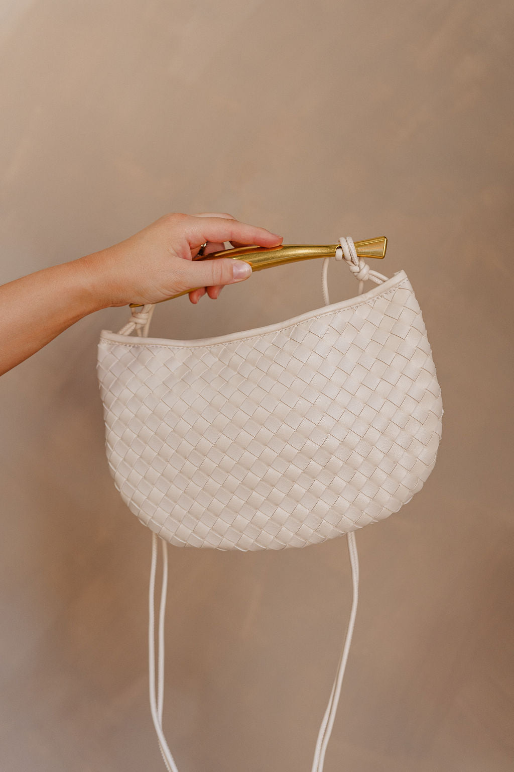 Hand is holding the Nadia Woven Purse on her shoulder. It features a cream woven body topped with a gold bar accent and cream straps.