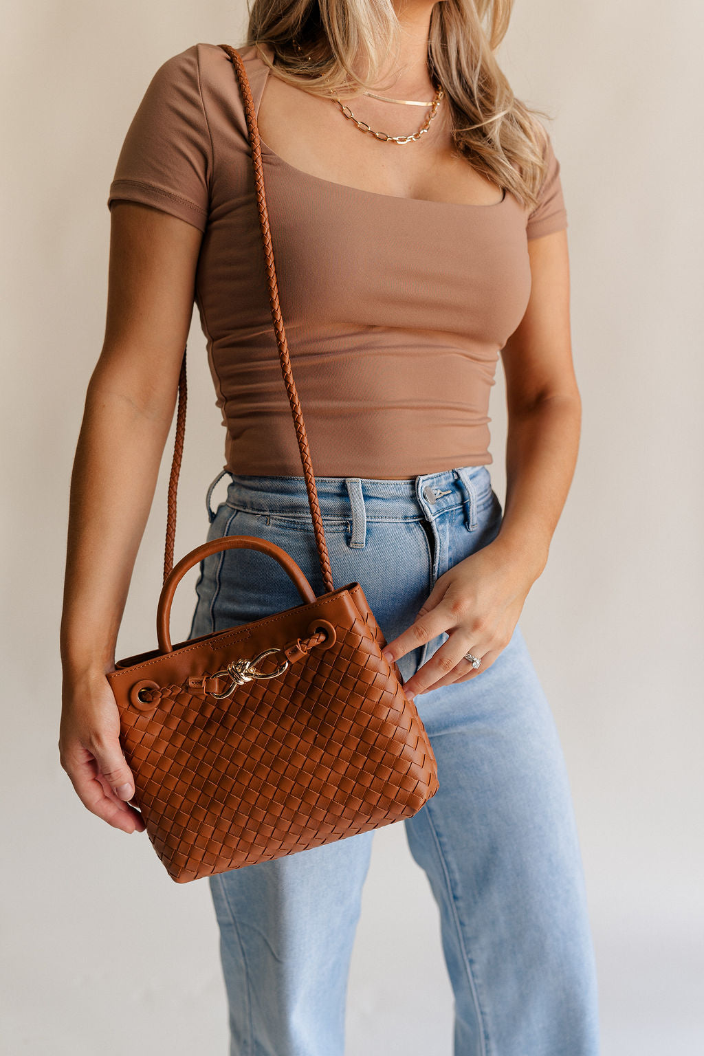 Female model is wearing the Blakely Light Brown Braided Purse that has a brown woven body, small handle, cross body strap, and a front gold accent detail. Shown on model's shoulder