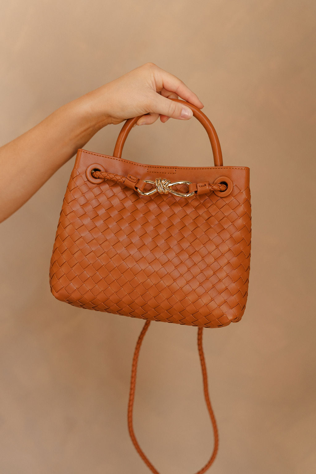 Model's hand is holding the Blakely Light Brown Braided Purse that has a brown woven body, small handle, cross body strap, and a front gold accent detail.