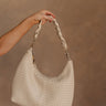 Hand is shown holding the Sabrina Braided Shoulder Tote in Cream that has a woven body, top zipper, and a braided shoulder strap.