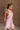Upper body side view of female model wearing the Eva Pink Sheer Tie Back Mini Dress that has pink sheer fabric with pink lining and a halter neck with a bow tie back