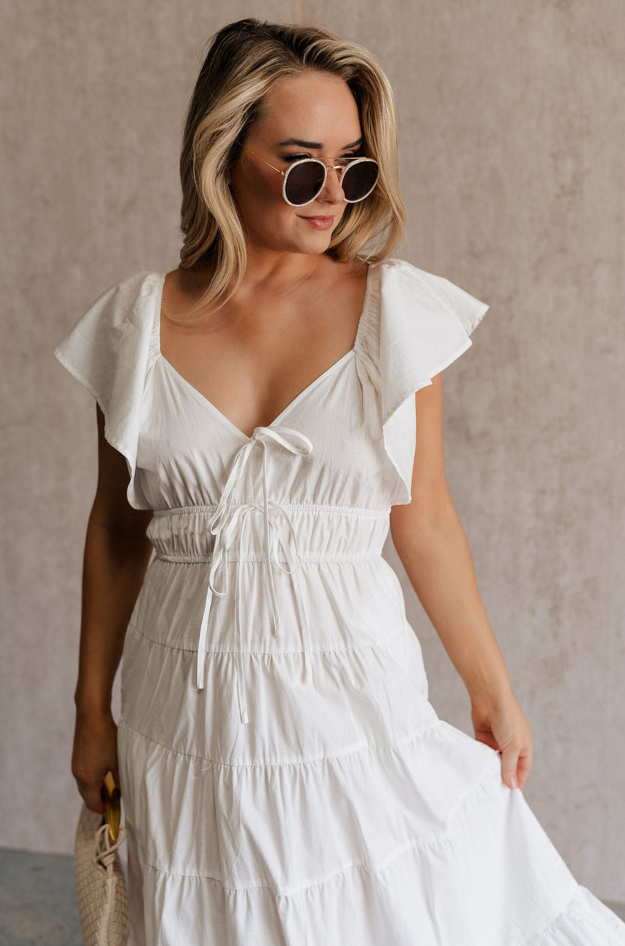 Front view of female model wearing the Gianna Ruffle Short Sleeve Tiered Maxi Dress in White which features Lightweight Fabric, Tiered Body, Midi Length, Pockets on each side, V-Neckline with a Tie Detail and Ruffle Short Sleeves. the dress is available in pink, aqua blue and white.