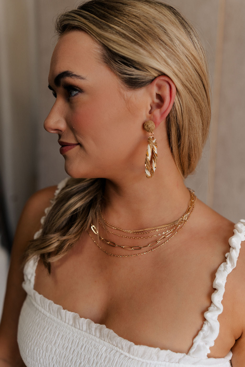 Side view of model's face; model is wearing the Nyomi Raffia & Gold Earrings that have ivory and tan raffia intertwined with gold metal into a circle hanging from tan raffia ball.