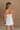 Upper body back view of female model wearing the Kelsey White Denim Romper that has white denim fabric, a strapless neck, and a pleated skirt over shorts lining.