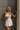 Upper body front view of female model wearing the Kelsey White Denim Romper that has white denim fabric, a strapless neck, and a pleated skirt over shorts lining. Worn with tan sling bag.