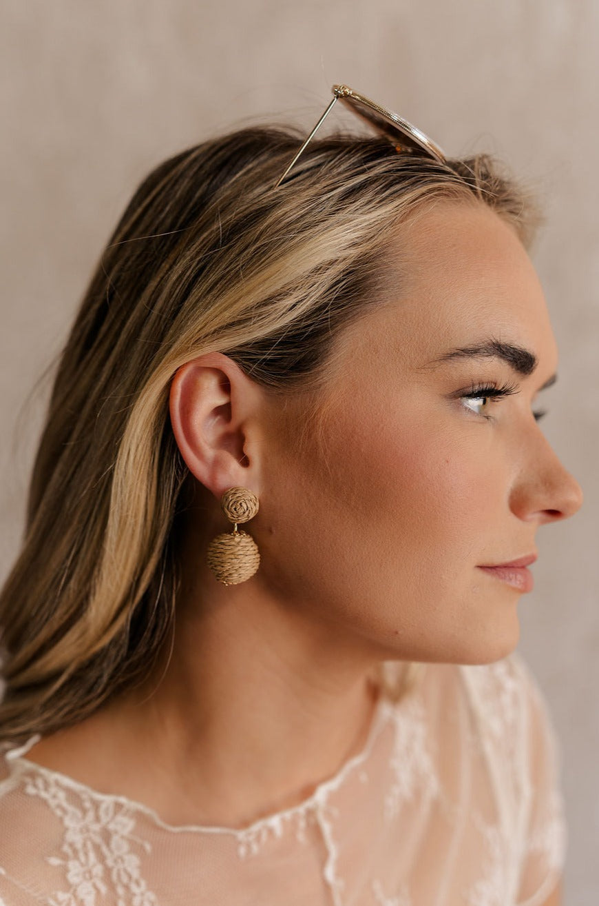 Side view of female model's face; model is wearing the Aubriella Raffia Lantern Earrings that have tan raffia balls dangling.