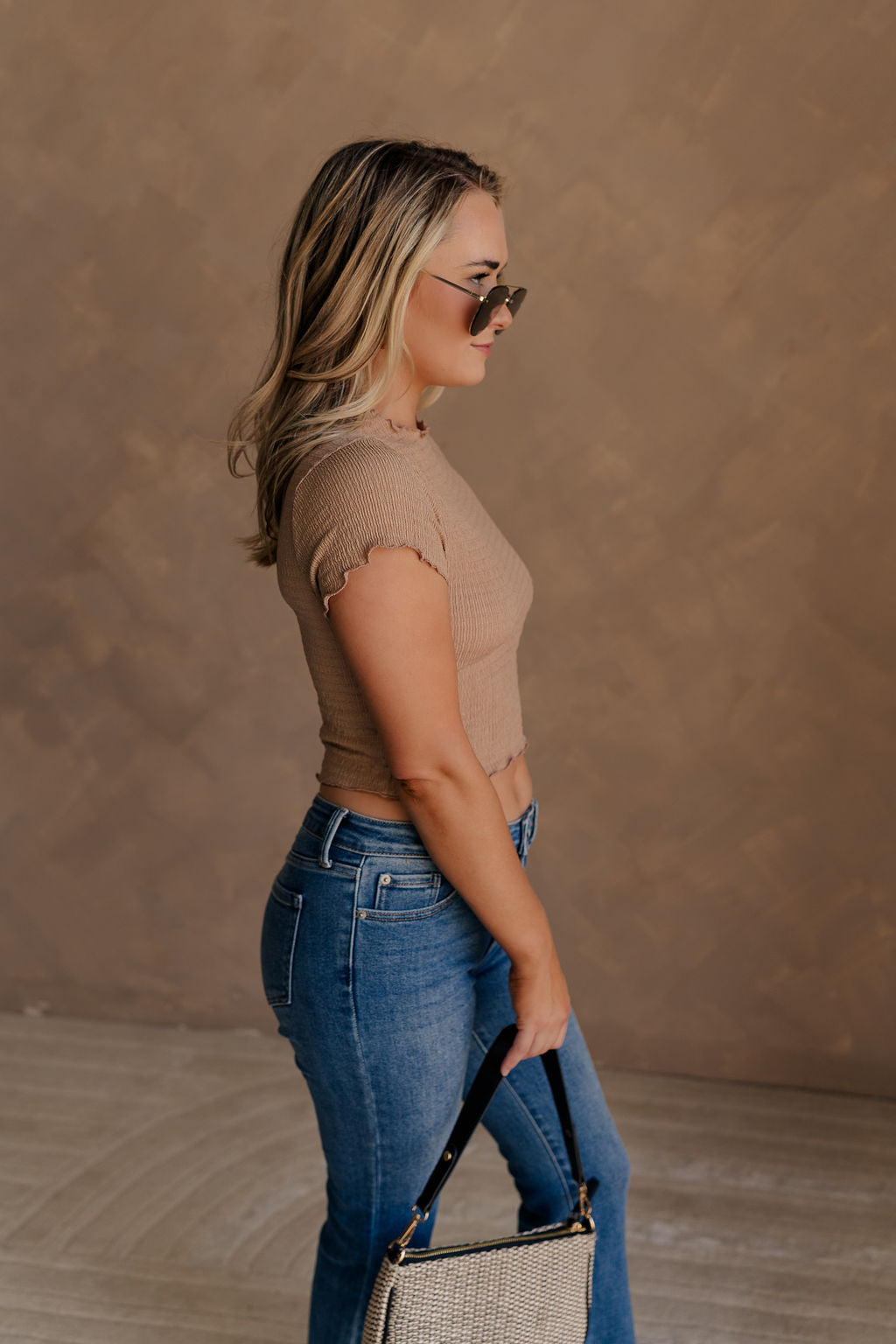 Side view of female model wearing the Nora Smocked Lettuce Hem Short Sleeve Top in tan that has stretchy textured fabric, lettuce trim, short sleeves, and a cropped fit.