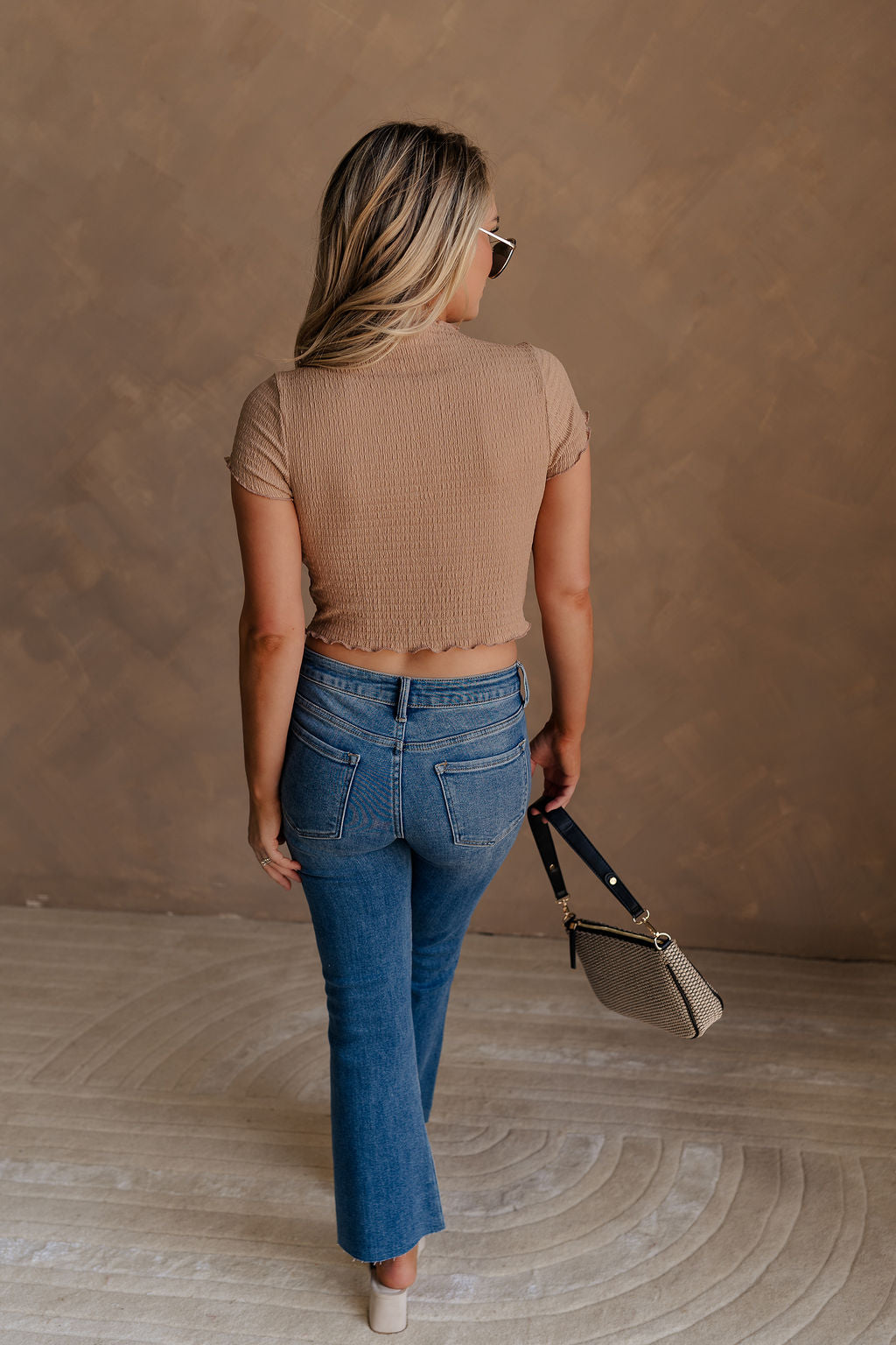 Full body back view of female model wearing the Nora Smocked Lettuce Hem Short Sleeve Top in tan that has stretchy textured fabric, lettuce trim, short sleeves, and a cropped fit.