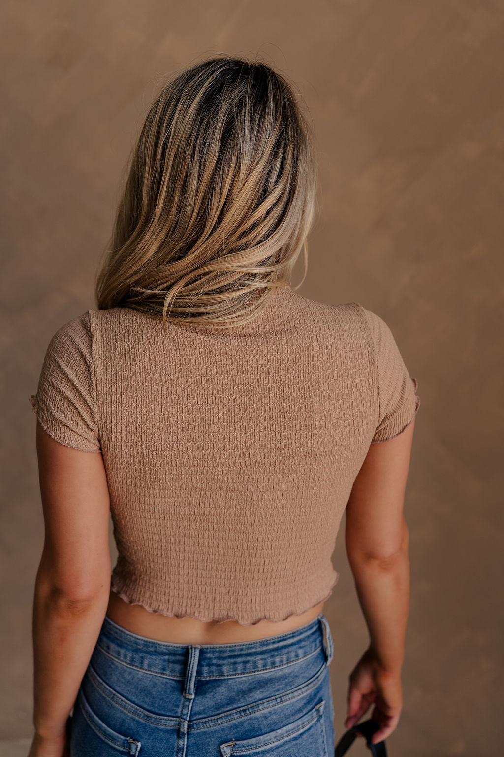 Back view of female model wearing the Nora Smocked Lettuce Hem Short Sleeve Top in tan that has stretchy textured fabric, lettuce trim, short sleeves, and a cropped fit.