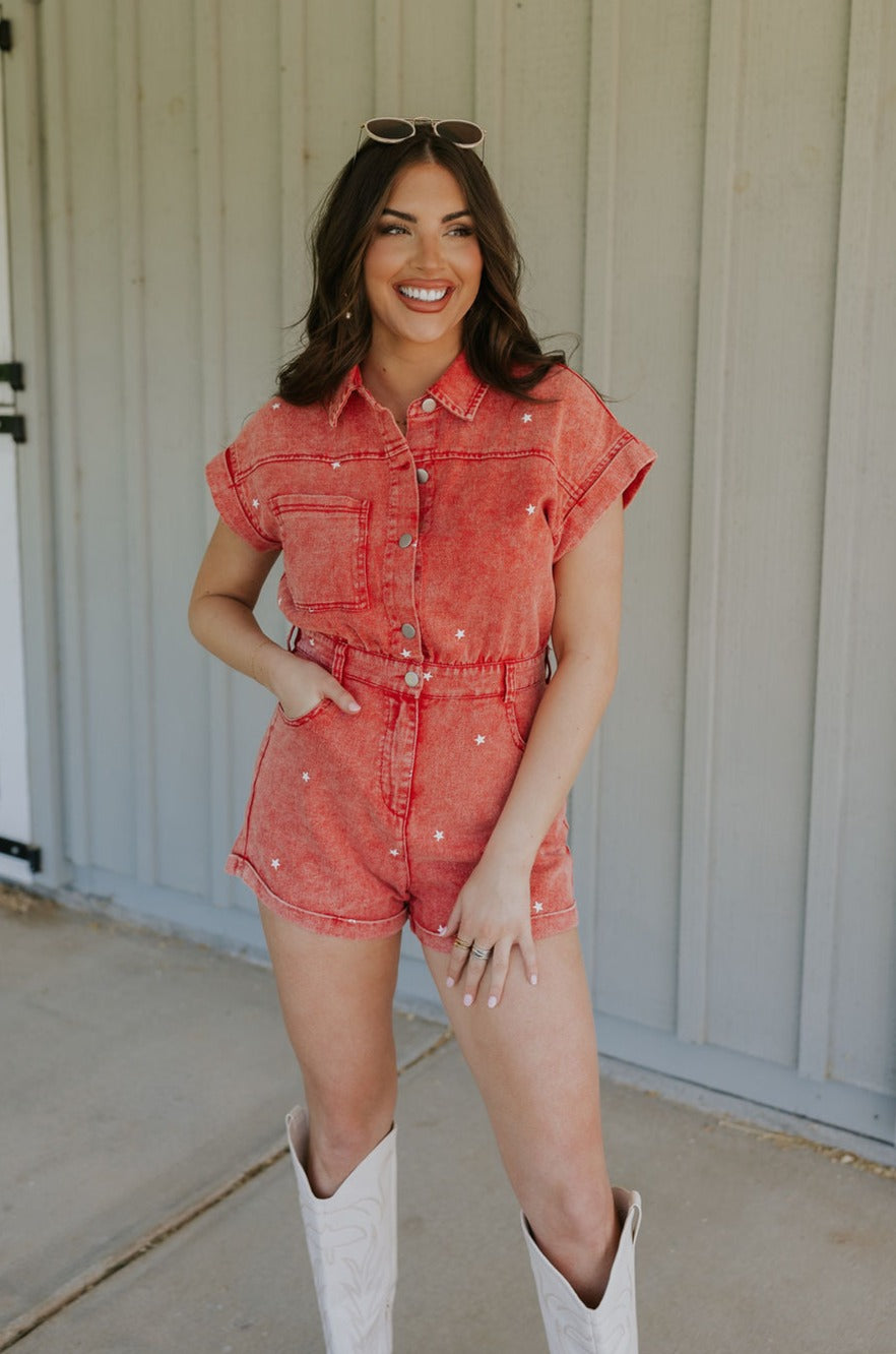 Front view of female model wearing the Krystal Washed Red Denim Star Romper that has red denim fabric with white stars, a snap up front, collar, belt loops, pocket, and short sleeves.