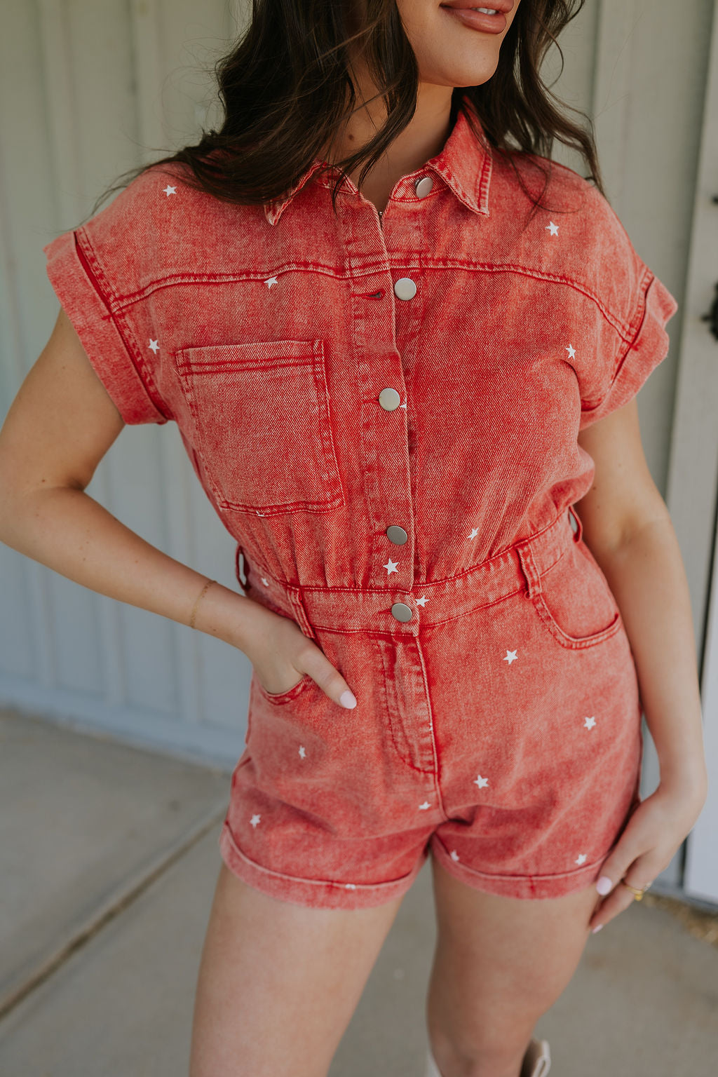 Close-up front view of female model wearing the Krystal Washed Red Denim Star Romper that has red denim fabric with white stars, a snap up front, collar, belt loops, pocket, and short sleeves.
