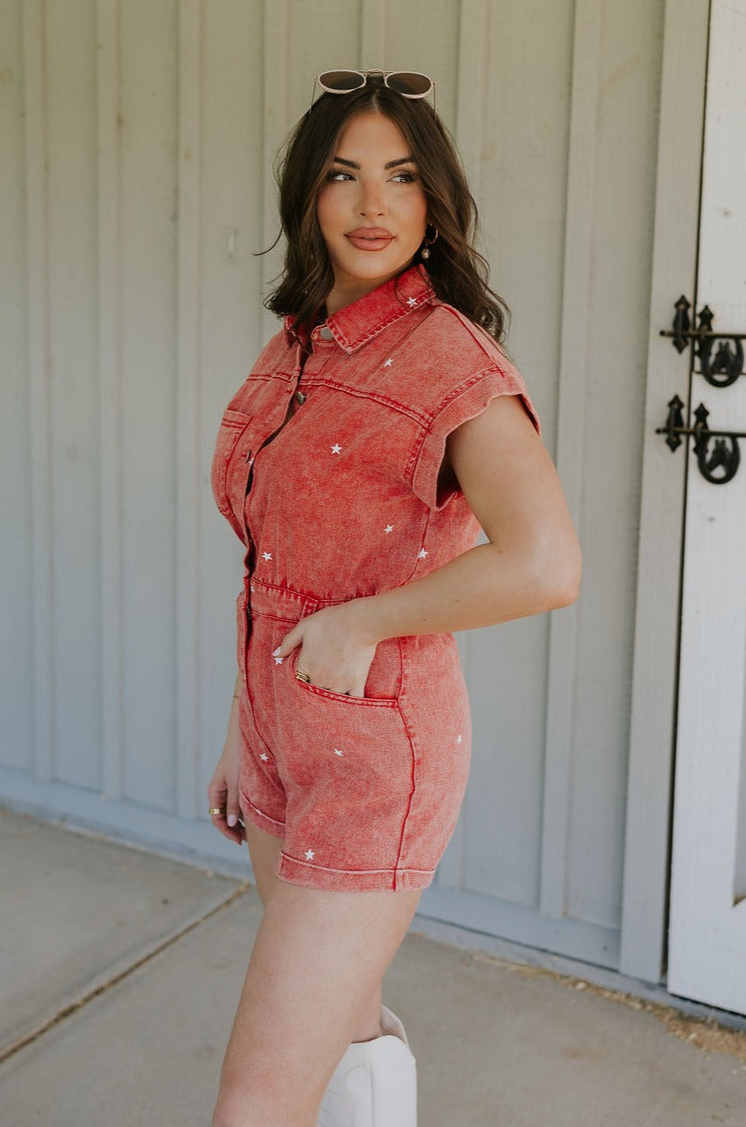 Side view of female model wearing the Krystal Washed Red Denim Star Romper that has red denim fabric with white stars, a snap up front, collar, belt loops, pocket, and short sleeves.