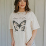 Upper body front view of female model wearing the Born To Fly Butterfly Graphic Tee that has cream fabric, a monarch butterfly graphic, "born to fly text", a round neck, oversized, and short sleeves.