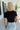 Upper body back view of blonde model wearing the Kira Short Sleeve Cropped Top in Black that has black fabric. short sleeves, a round neck, and cropped waist. Worn with denim shorts.