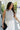 Front view of female model wearing the Lily White & Black Striped Tank Top that has white textured fabric with black vertical stripes, a high round neck, and sleeveless body. Top is tucked into white pants.