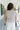 Upper body back view of female model wearing the Lily White & Black Striped Tank Top that has white textured fabric with black vertical stripes, a high round neck, and sleeveless body.