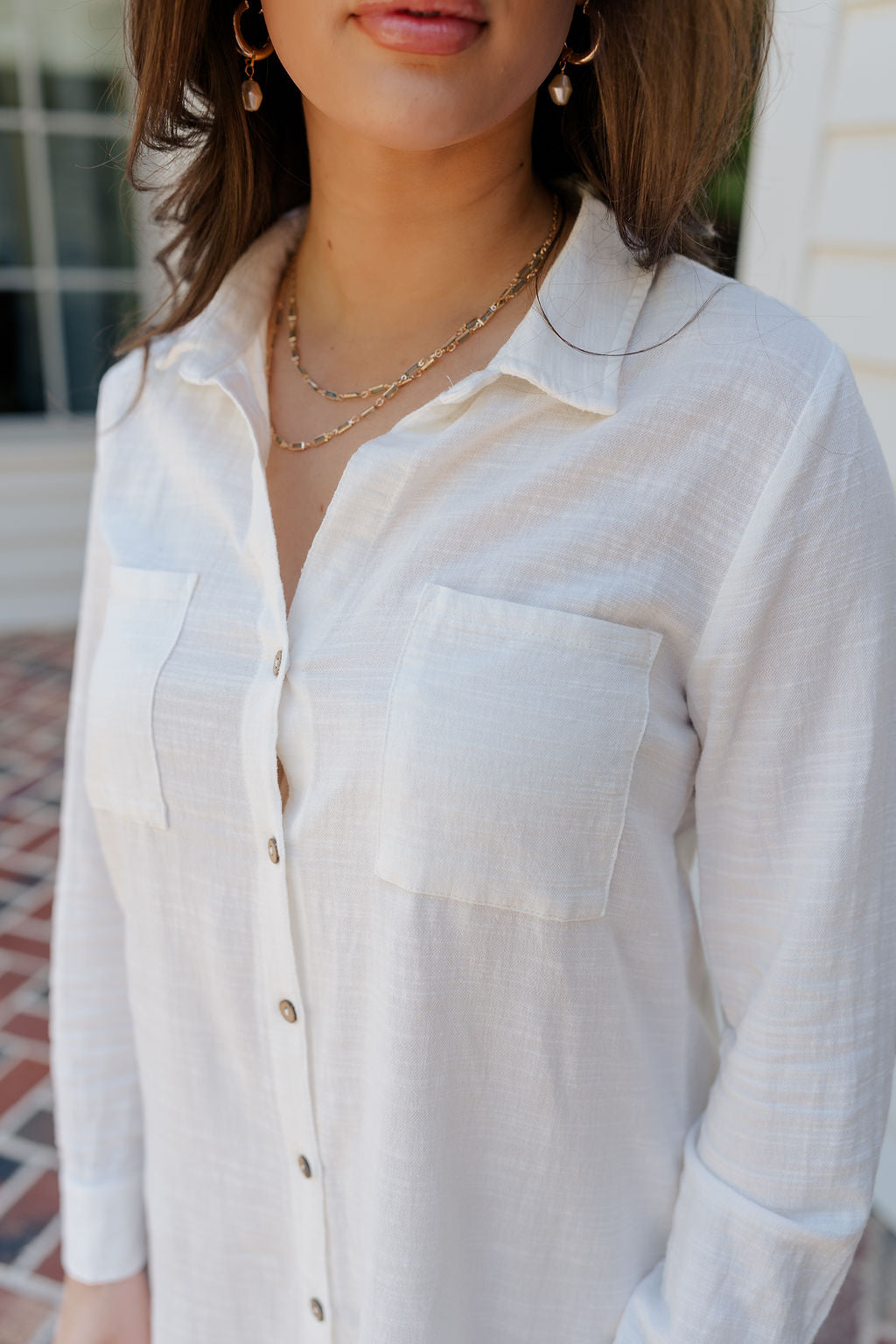 Close-up side frontal view of female model wearing the Everly White Button Up Midi Dress that has white fabric, long sleeves, a button up front, collar, chest pockets, and a frayed high-low hemline with back slit.