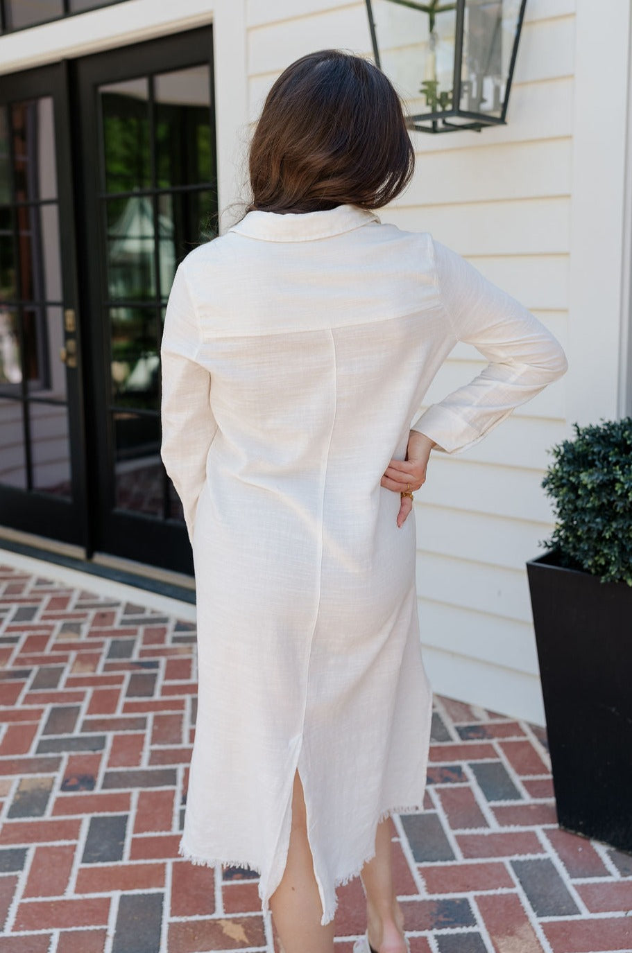 Back view of female model wearing the Everly White Button Up Midi Dress that has white fabric, long sleeves, a button up front, collar, chest pockets, and a frayed high-low hemline with back slit.