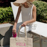 Front view of model holding the Hola Beaches Natural Tote which features lightweight natural woven fabric, two sturdy straps and pink stitching "hola beaches."