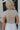 Upper body back view of blonde model wearing the Kira Short Sleeve Cropped Top in Fog that has grey fabric, short sleeves, a round neck, and cropped waist. Worn with white pants.