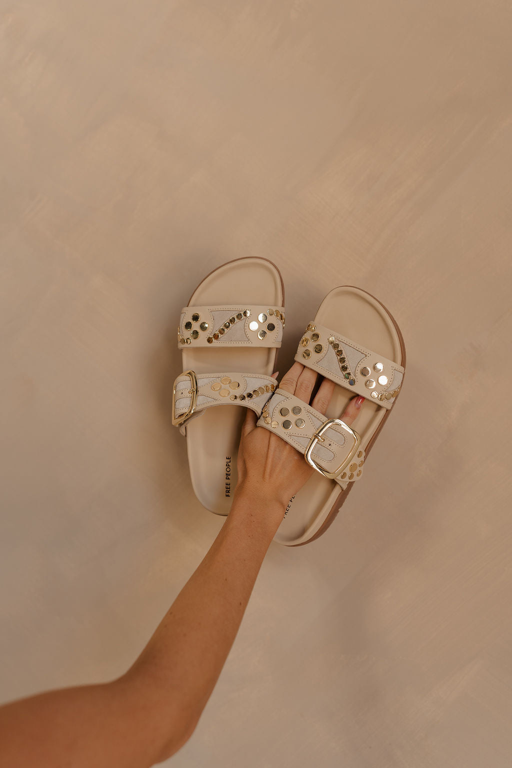 Front view of female model holding the Revelry Studded Sandal which features light taupe suede upper, gold stud details, double strap, adjustable buckle closure and slide-on style