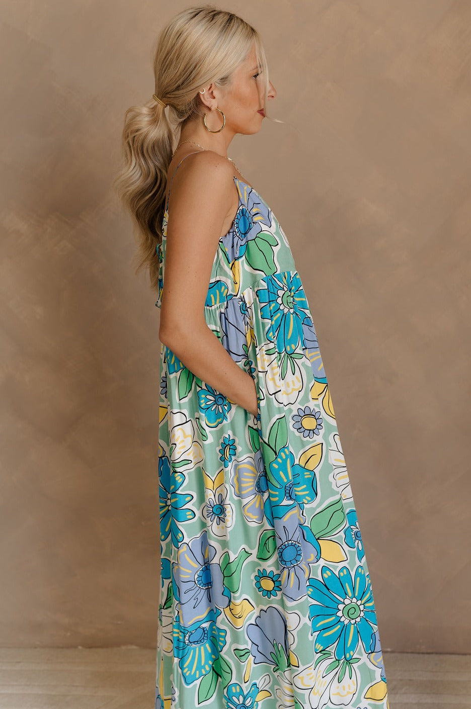 Side view of female model wearing the Mallory Green Multi Floral Sleeveless Midi Dress which features Green, blue, white, and yellow floral print, Round neckline, Adjustable straps, Keyhole back with tie closure and White lining