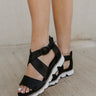 Side view of female model wearing the Mackenna Sandal in Black which features smooth, sleak black leather upper, white rugged sole, adjustable buckle, crisscross straps, cushioned footbed and back zipper with leather pull