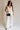 Full body view of female model wearing the Amelia Off White & Black Wide Leg Pants which features  Off White Linen Fabric, Black Trim Details, Front Pockets, Wide Pant Legs, Front Zipper with Tortoise Button Closure and Belt Loops