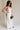 Full body side view of female model wearing the Amelia Off White & Black Wide Leg Pants which features Off White Linen Fabric, Black Trim Details, Front Pockets, Wide Pant Legs, Front Zipper with Tortoise Button Closure and Belt Loops