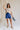 Full body front view of model wearing the Mara Blue High Waist Shorts that have blue fabric, a high-rise elastic back waist. Worn with beige top.