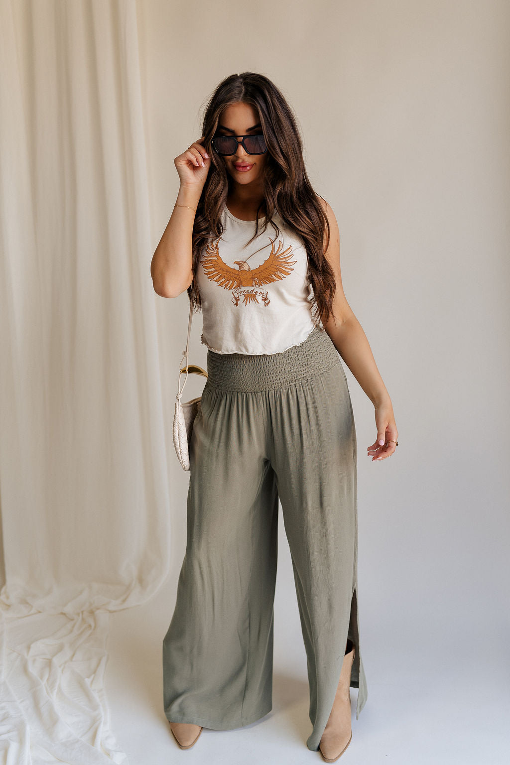 Full body front view of female model wearing the Audrey Olive Green Wide Leg Pants that have olive green fabric, a smocked waist, wide legs, and side slits. Worn with beige tank top