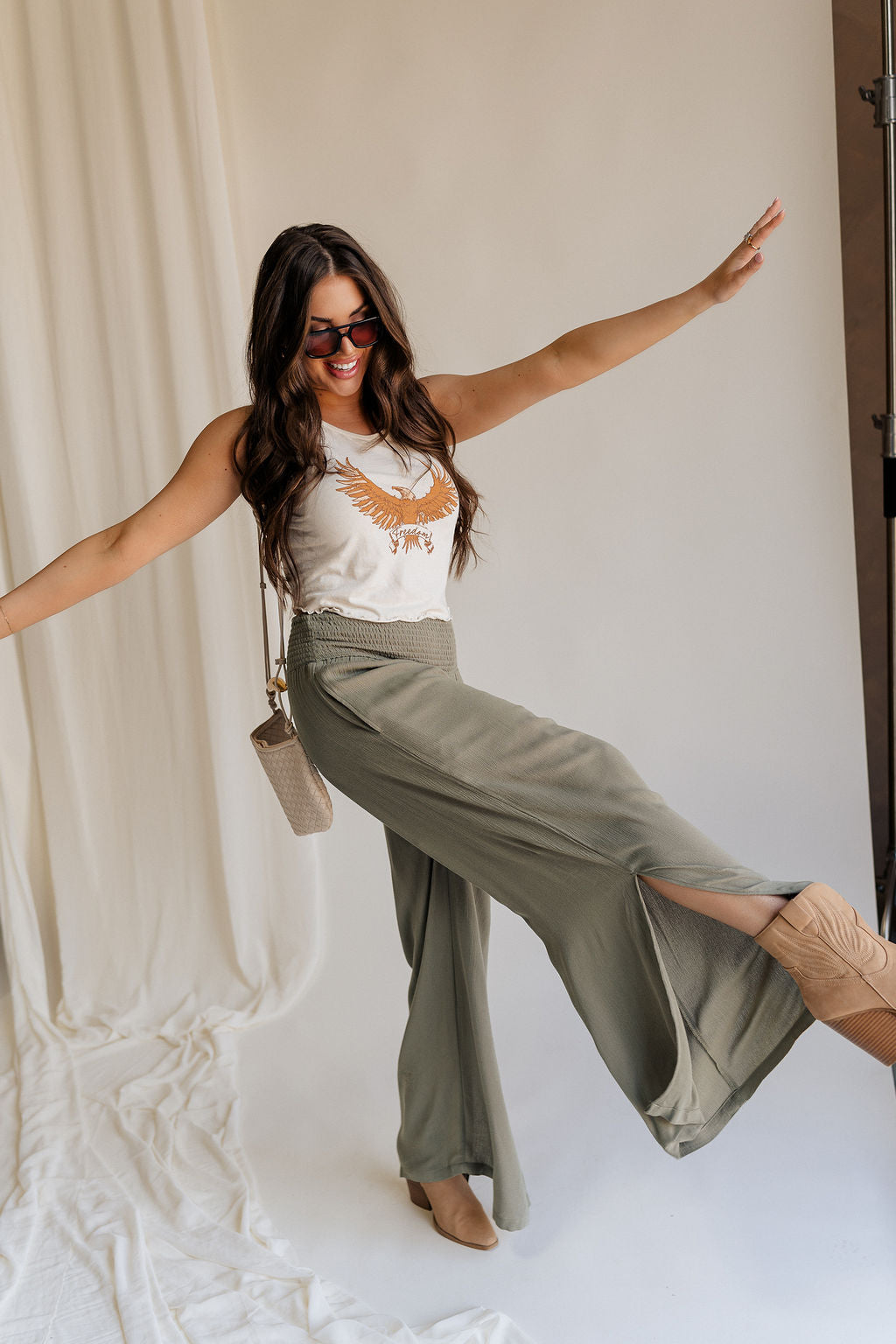 side view of female model wearing the Audrey Olive Green Wide Leg Pants that have olive green fabric, a smocked waist, wide legs, and side slits. Worn with beige tank top. Model is kicking out leg.