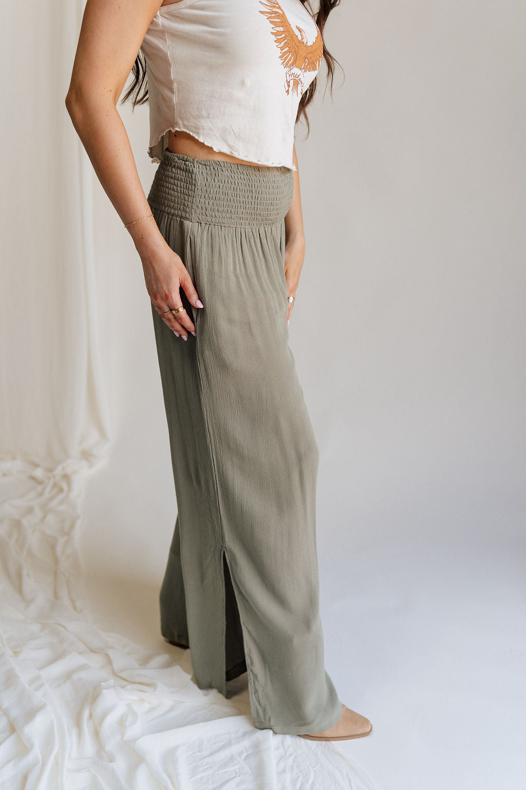 Lower body side  view of female model wearing the Audrey Olive Green Wide Leg Pants that have olive green fabric, a smocked waist, wide legs, and side slits. Worn with beige tank top
