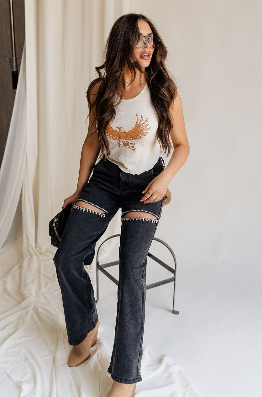 Full body front view of female model wearing the Remi Rhinestone Slit Black Jeans that have black denim, thigh slits with rhinestone trim, and pockets. Model is sitting on stool.