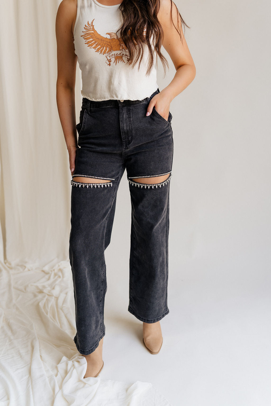 Lower body front view of female model wearing the Remi Rhinestone Slit Black Jeans that have black denim, thigh slits with rhinestone trim, and pockets.