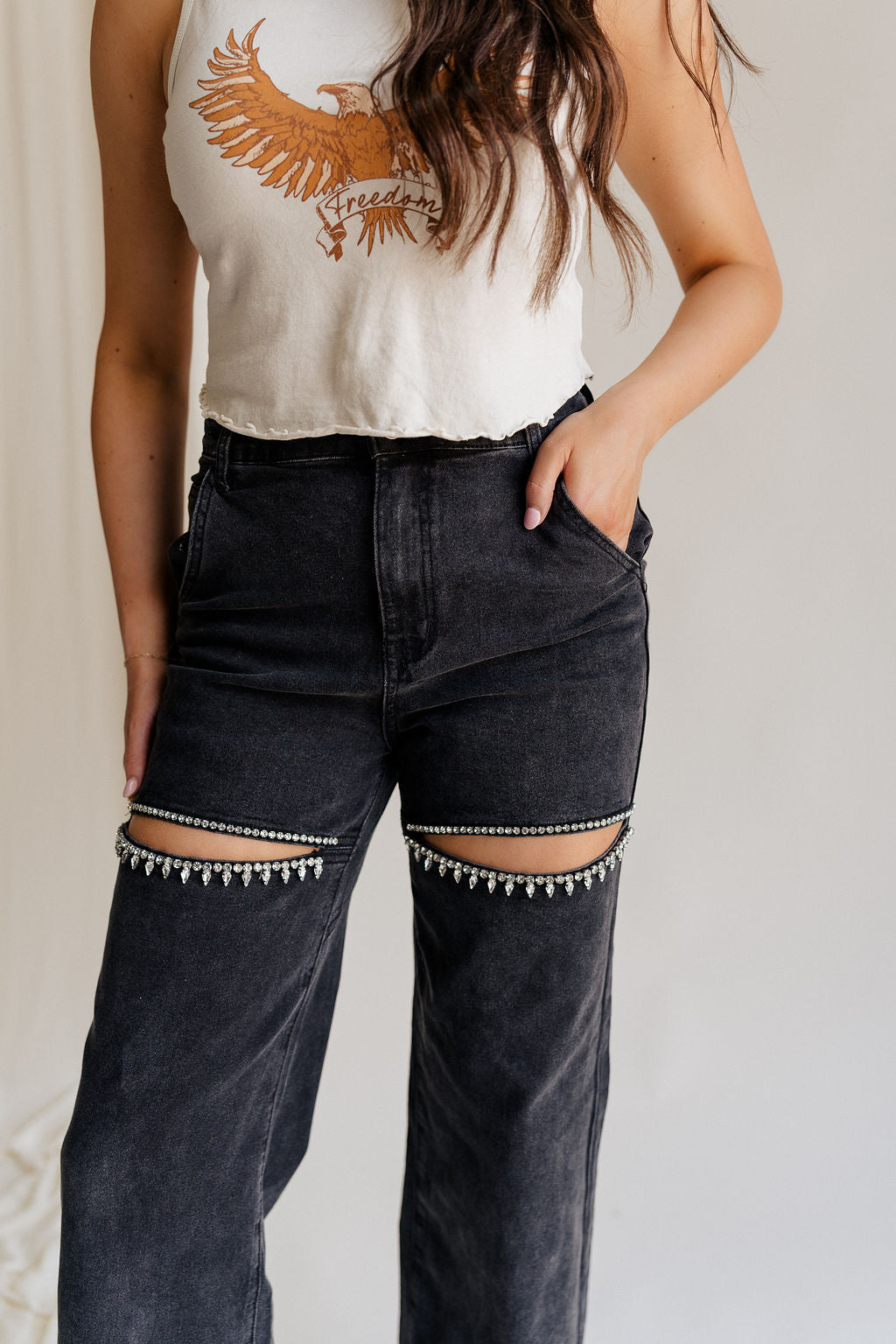 Close up front view of female model wearing the Remi Rhinestone Slit Black Jeans that have black denim, thigh slits with rhinestone trim, and pockets.