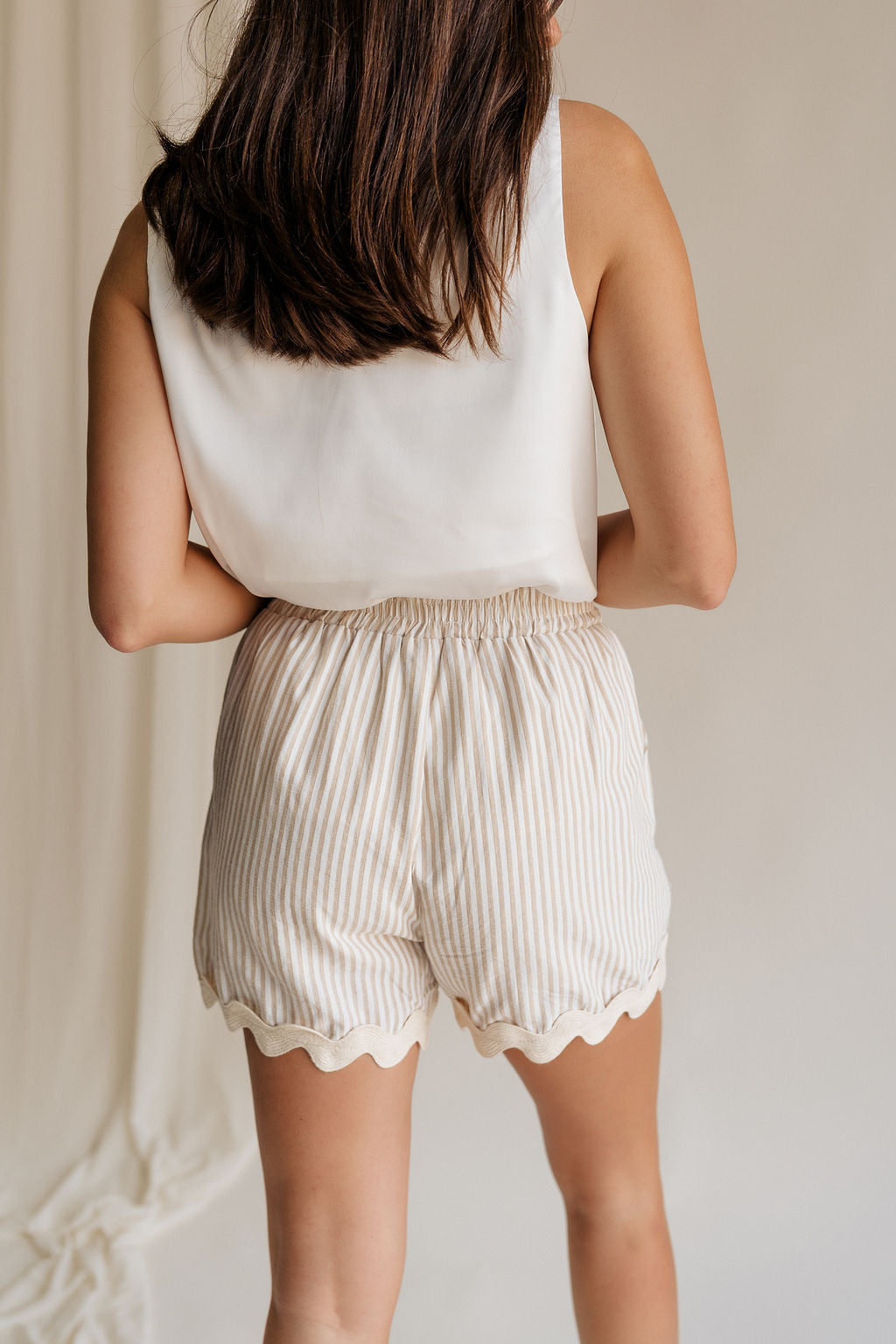 back view of female model wearing the Patricia Beige & Cream Stripe Scallop Hem Shorts which features Beige and White Stripe Print, White Lining, Scalloped Hem, Elastic Waistband and Side Pockets