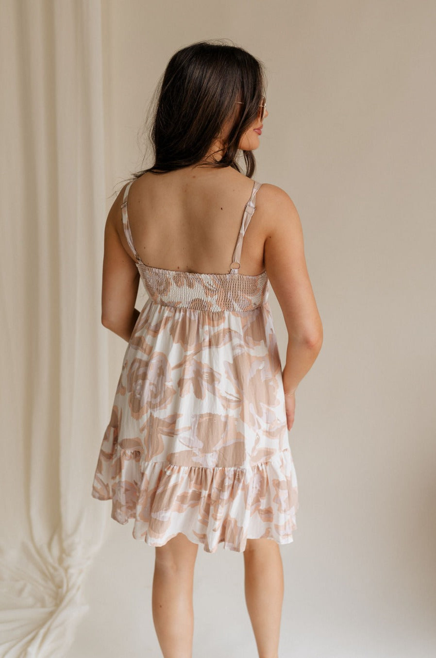 Back view of female model wearing the Shelby Floral Sleeveless Mini Dress that has ivory fabric with peach and taupe floral print, sleeveless body, and a straight neckline.