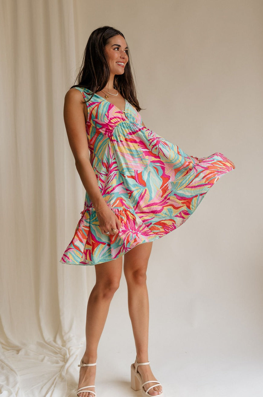 Full body front view of female model wearing the Florence Floral Print Sleeveless Mini Dress that has a colorful floral palm print, v neck and back, and mini length.