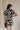 Back view of model wearing the Melanie Black & Cream Printed Romper that has black and cream fabric with a swirl print, a button up front, collar, tie belt, and short sleeves.