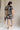 Full body back view of model wearing the Melanie Black & Cream Printed Romper that has black and cream fabric with a swirl print, a button up front, collar, tie belt, and short sleeves.
