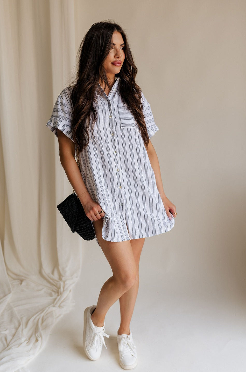 Full body front view of female model wearing the Jenna White & Black Striped Mini Dress that has white fabric with vertical black stripes, a front pocket, button up front, short sleeves, and collar.
