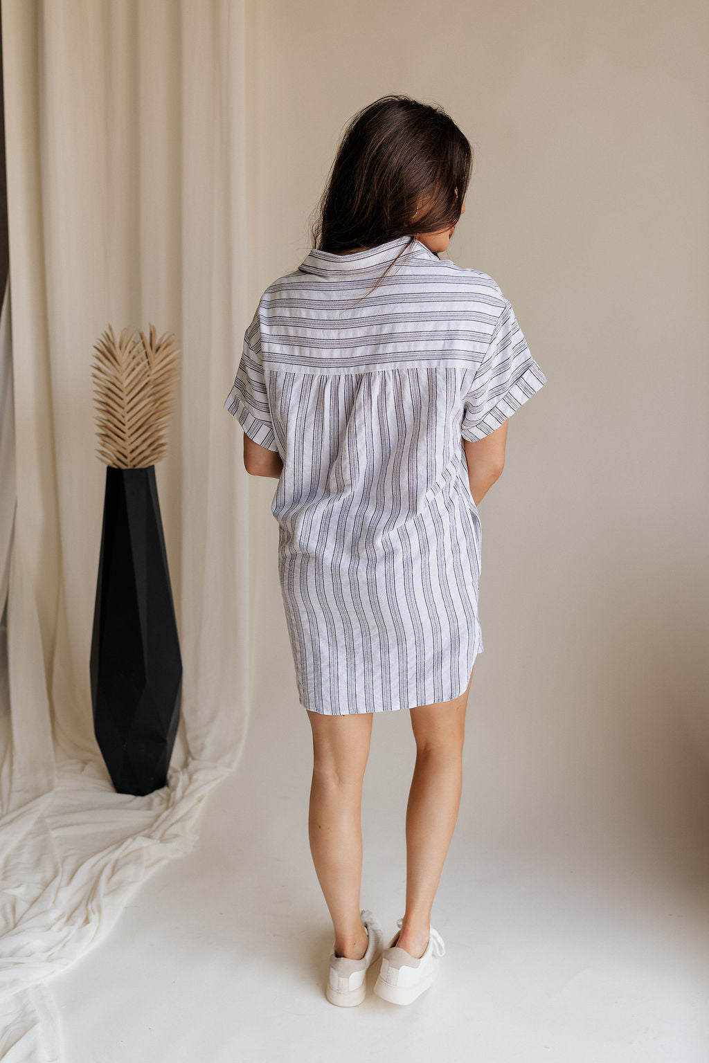 Full body back view of female model wearing the Jenna White & Black Striped Mini Dress that has white fabric with vertical black stripes, a front pocket, button up front, short sleeves, and collar.