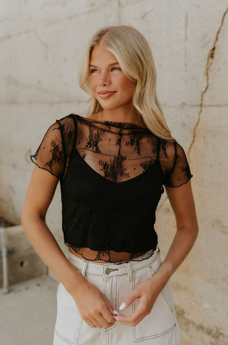 Upper body front view of female model wearing the Vanessa Cropped Lace Top in Black that has a short sleeve sheer lace top over an ivory cami. Worn with white denim skirt.