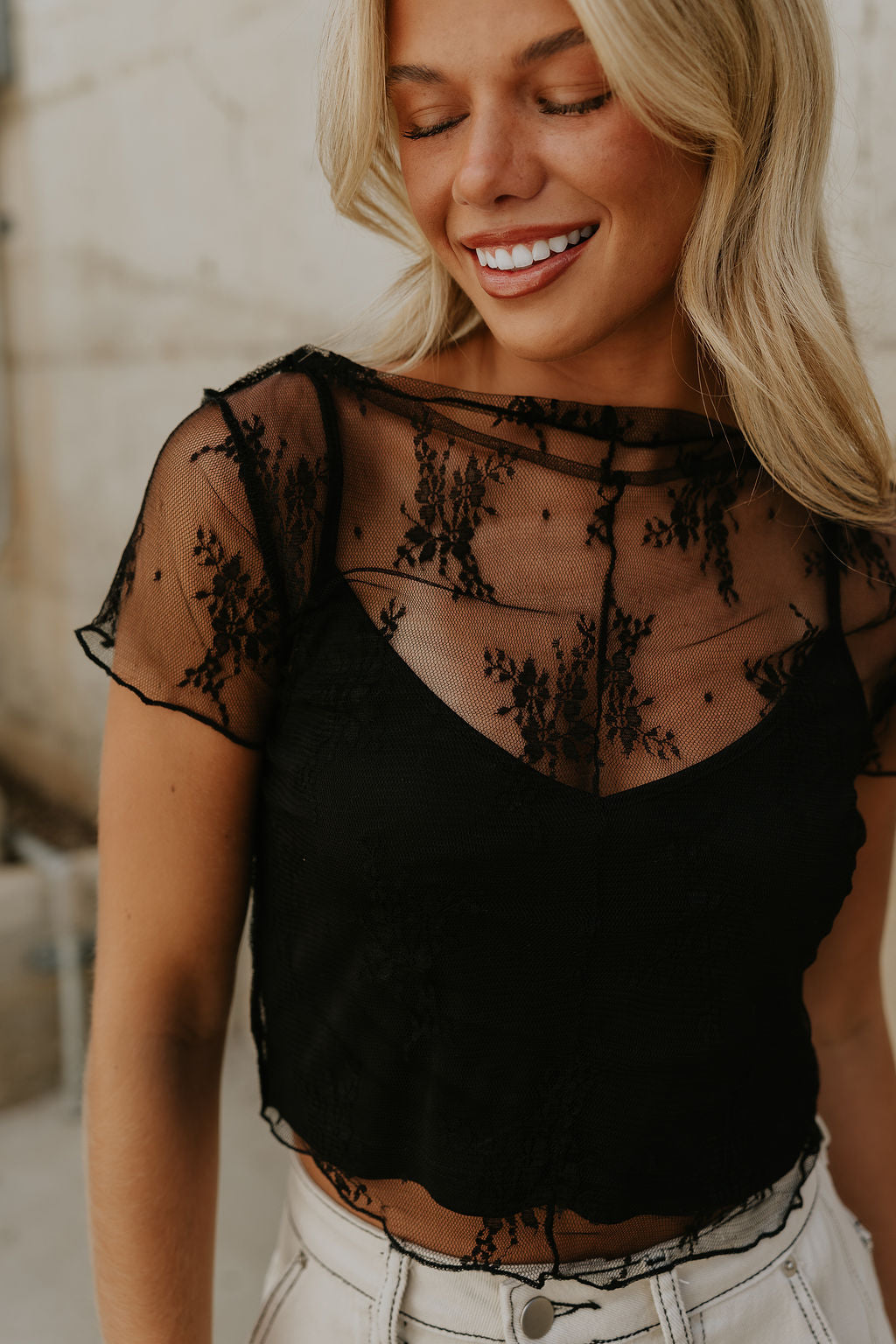 Close-up front view of female model wearing the Vanessa Cropped Lace Top in Black that has a short sleeve sheer lace top over an ivory cami. Worn with white denim skirt.