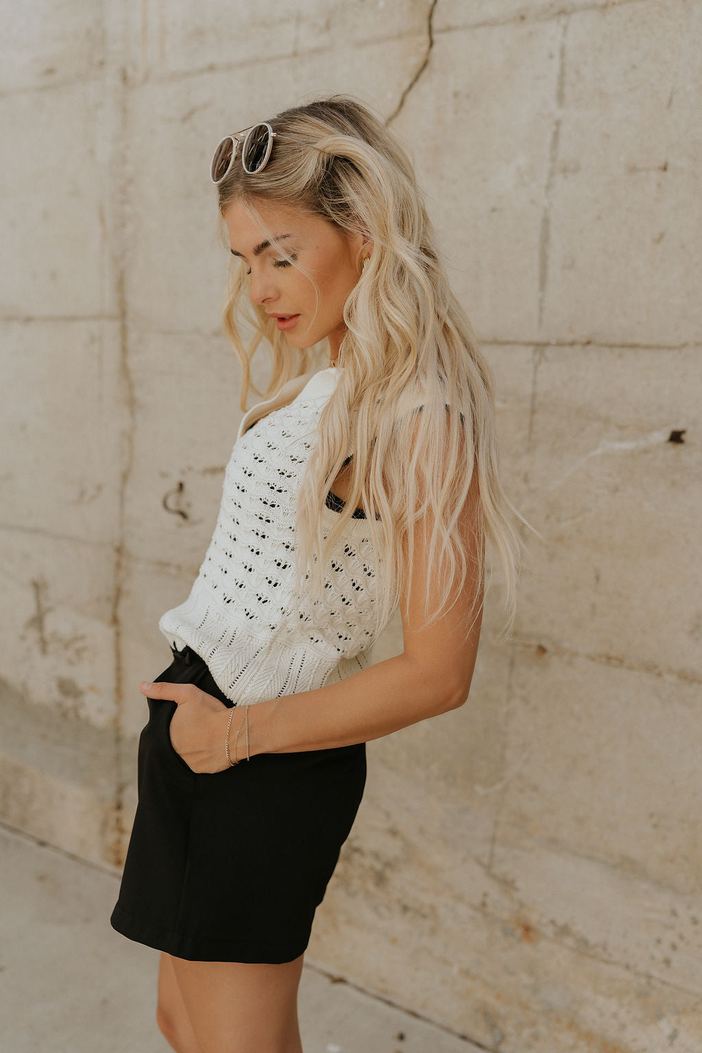 Upper body side view of female model wearing the Ariana Cream Crochet Tank Top that has cream crochet fabric, black trim, scalloped hem, and a collar. Worn with black shorts.